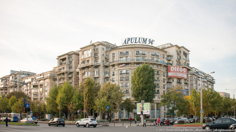 Large curved apartment complex at a Bucharest intersection, with rooftop “APULUM 94” lettering, trees, shops, and traffic.