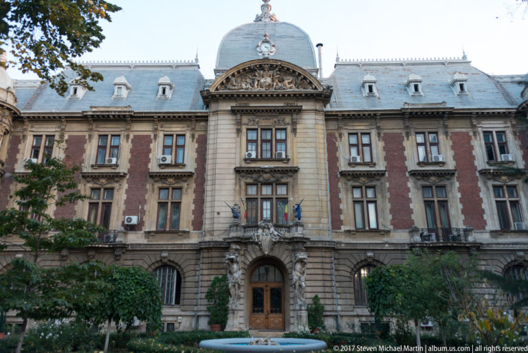 Symmetrical historic building with rusticated stonework, tall windows, slate roof, dormers, central pediment, and Romanian flags.