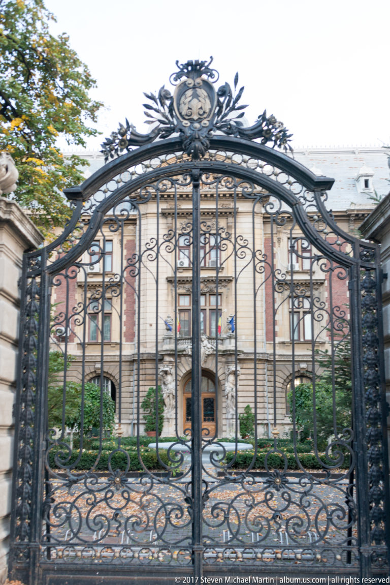 Black wrought-iron gate with scrolling ornament and a crest, framing a pale institutional building in Bucharest.