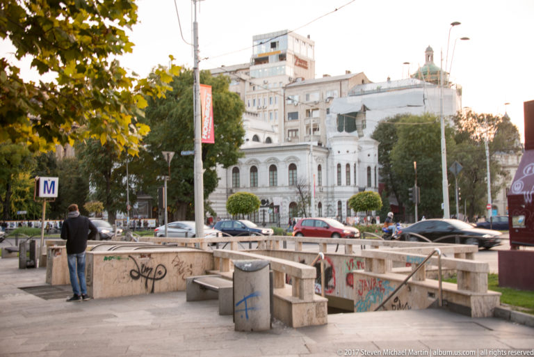 Sunken Bucharest metro entrance with concrete railings, benches, graffiti, traffic, trees, and a white historic building beyond.