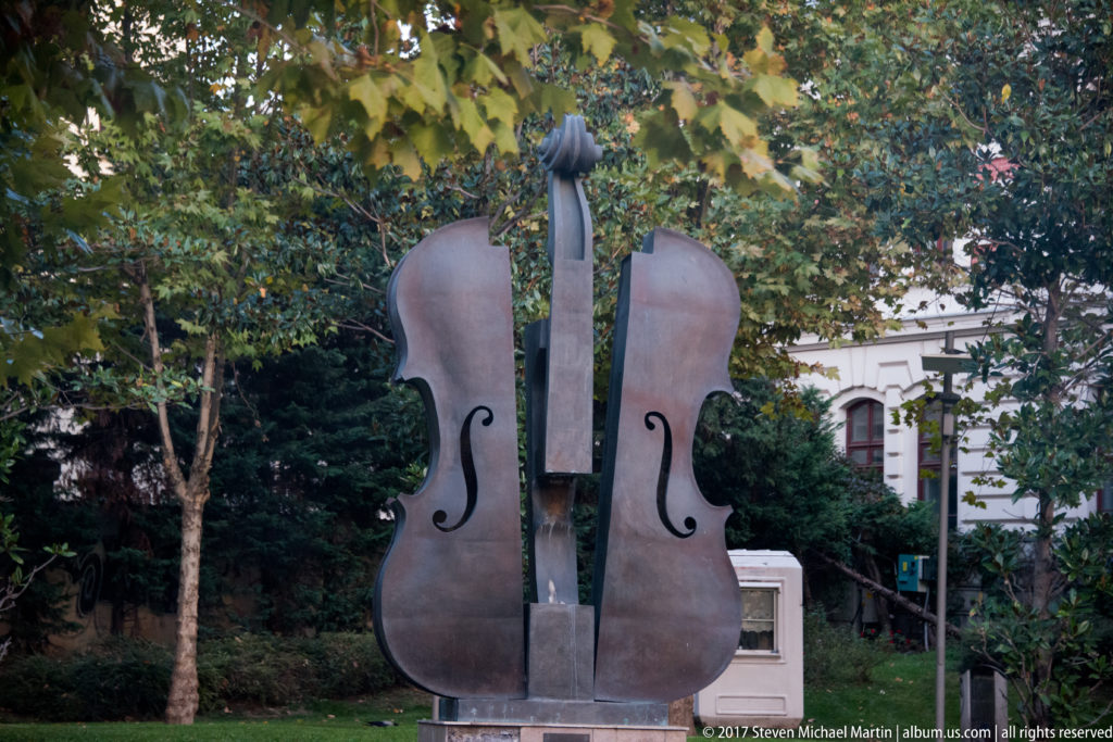 Large upright cello sculpture with dark f-holes and tall central neck standing among trees in a landscaped Bucharest setting.