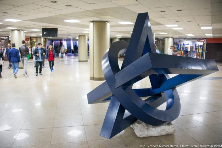 Large dark abstract metal sculpture of interlocking angular loops in a bright tiled Bucharest concourse with pedestrians behind.