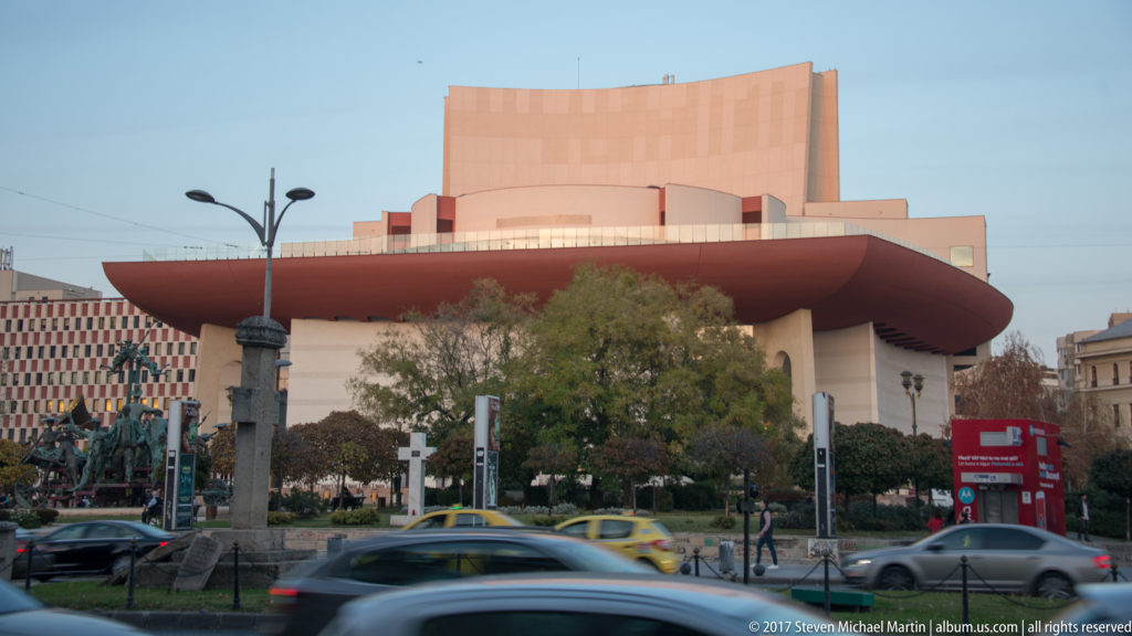 Ion Luca Caragiale National Theatre in Bucharest with tall warm-toned stage block, broad curved canopy, trees, and traffic in front.