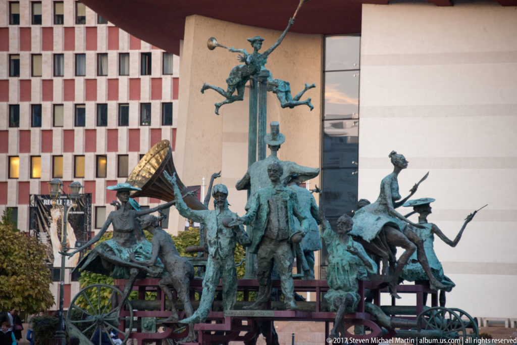 Caragialiana bronze sculpture ensemble with clustered theatrical figures, carriage elements, and performers outside Bucharest National Theatre.