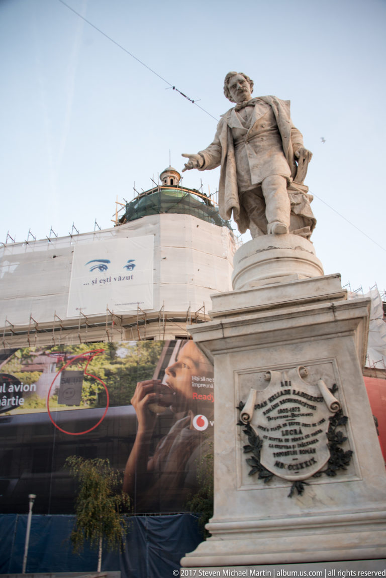 Spiru Haret statue gesturing from an ornate inscribed pedestal in University Square, with construction sheeting behind.