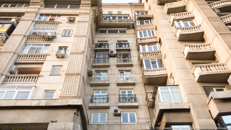 Upward view between tall Bucharest apartment blocks with stacked balconies, enclosed windows, air conditioners, and ribbed façades.