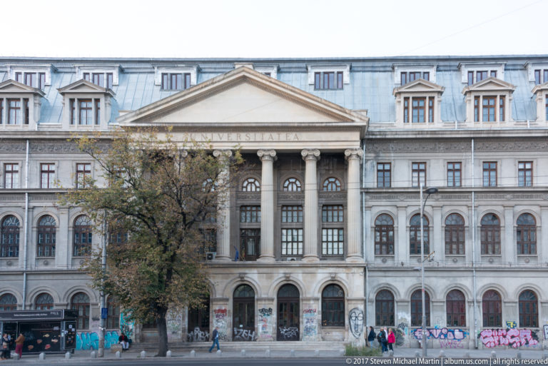 University of Bucharest façade with central columned portico, triangular pediment, rows of windows, graffiti, trees, and pedestrians.