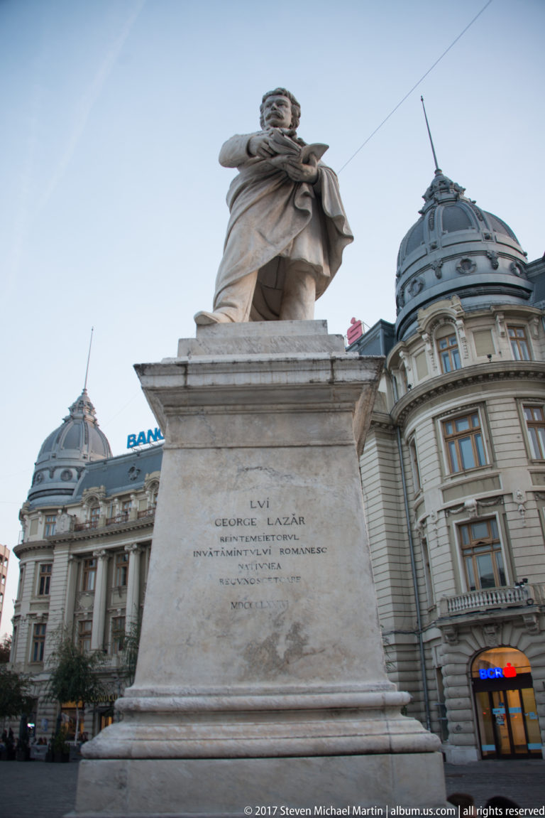 Gheorghe Lazăr statue on a tall inscribed pedestal in University Square, with ornate domed Bucharest buildings behind.