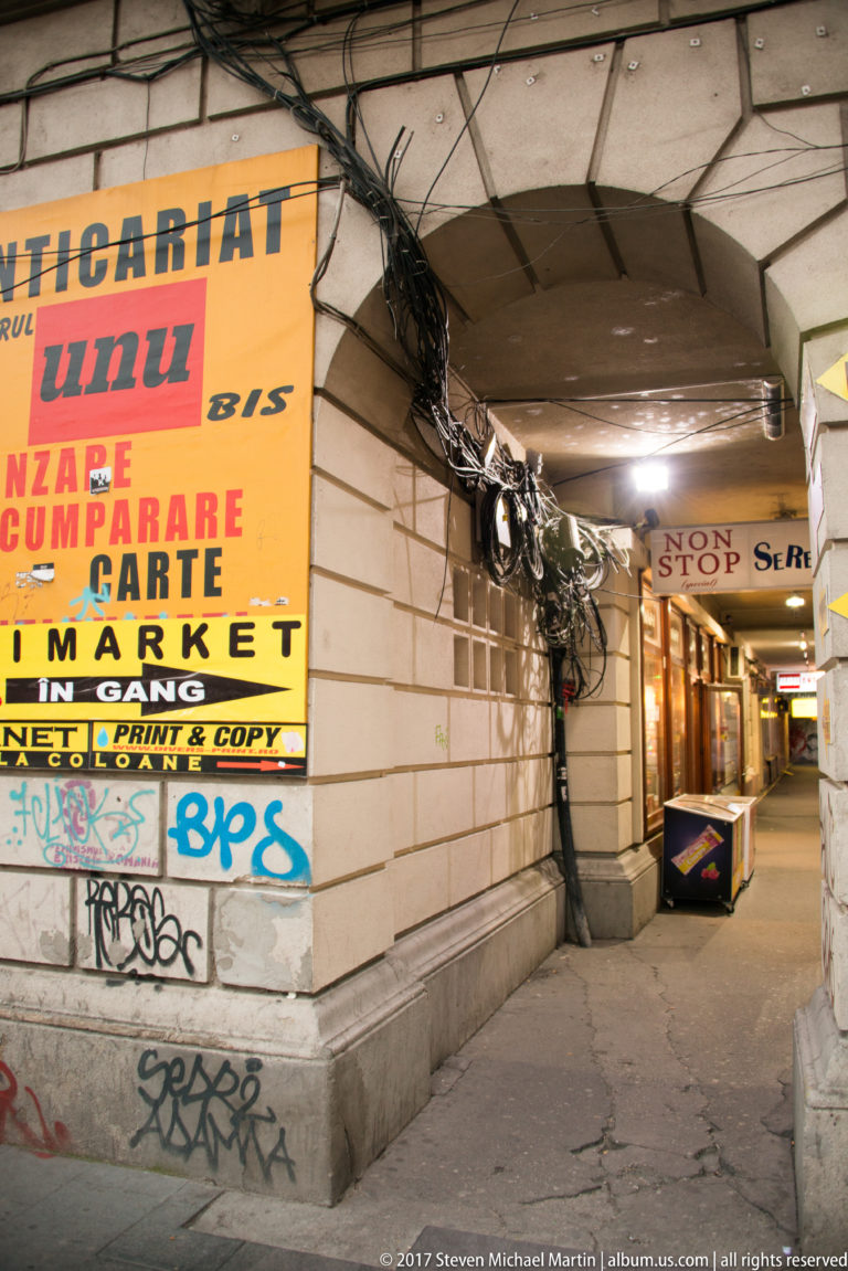 Low covered Bucharest walkway with restaurant and market signs, exposed cable bundles, graffiti, and small commercial entrances.