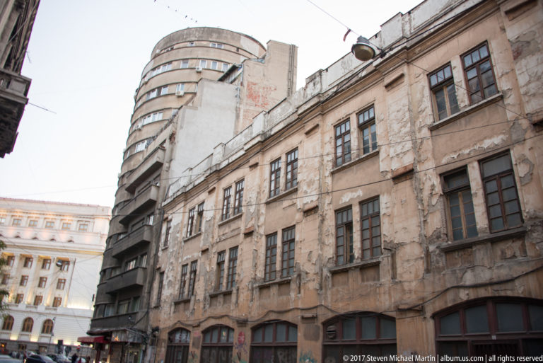 Weathered multi-story Bucharest building with tall windows in front of a rounded modernist structure, showing cracked and stained masonry.
