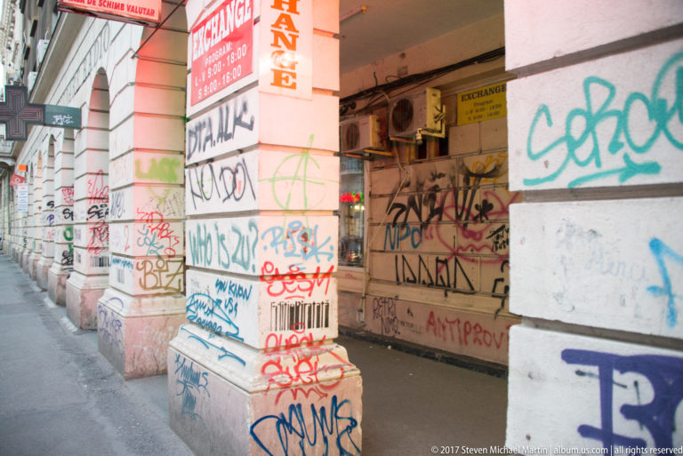 Sheltered Bucharest sidewalk with graffiti-covered columns and walls beside open shops and a currency-exchange storefront.