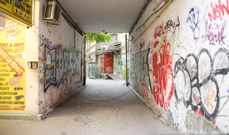 Narrow Bucharest passage with graffiti-covered walls, exposed utilities, air-conditioning units, and daylight opening into a rear court.