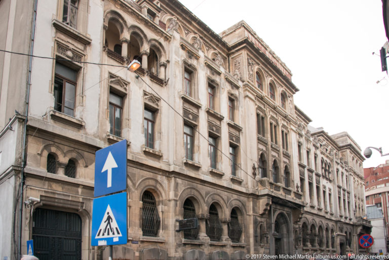 Weathered ornate historic building on a Bucharest street with arched windows, carved stone details, overhead wires, and pedestrian sign.