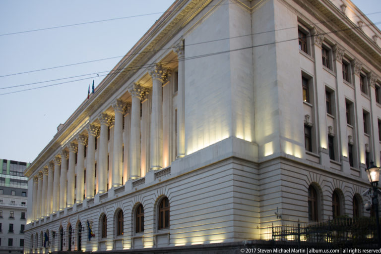 Long illuminated white neoclassical building in Bucharest with tall colonnade, gilded capitals, arched ground-floor openings, and Romanian flag.