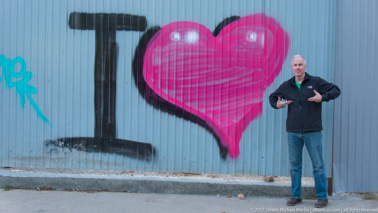 Man posing and pointing beside a large pink heart graffiti mural painted on a corrugated metal wall.