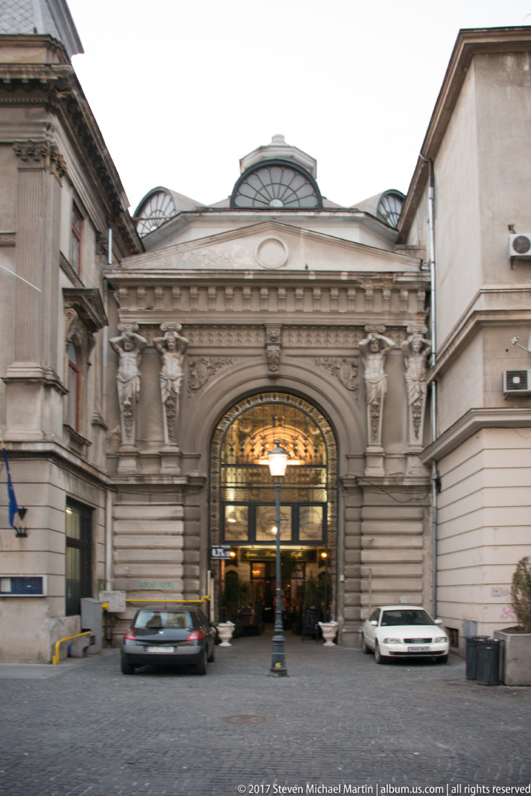 Full view of an ornate arched Bucharest passage entrance between buildings, with sculpted figures, roof glazing, parked cars, and bins.