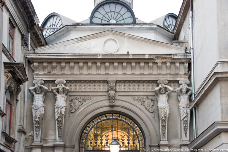 Ornate Bucharest arcade entrance with four sculpted female figures, rusticated stone arch, decorative iron doors, and semicircular roof windows.