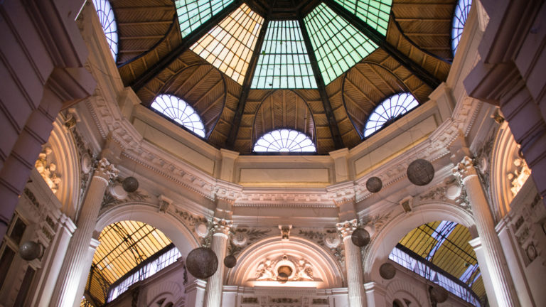 Upward view inside a Bucharest arcade rotunda with colored glass roof panels, fan windows, ornate arches, and hanging spherical lamps.