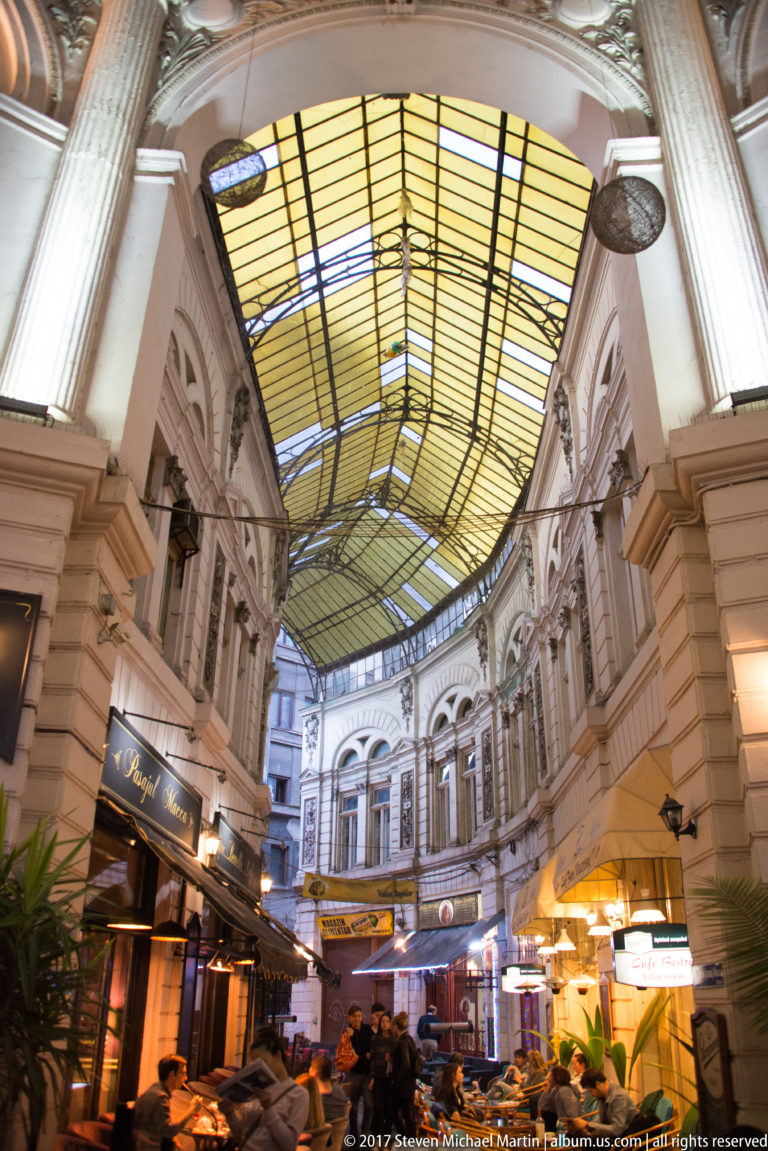 Long covered Bucharest arcade with glazed barrel roof, ornate façades, restaurant signs, café seating, and a decorated end wall.
