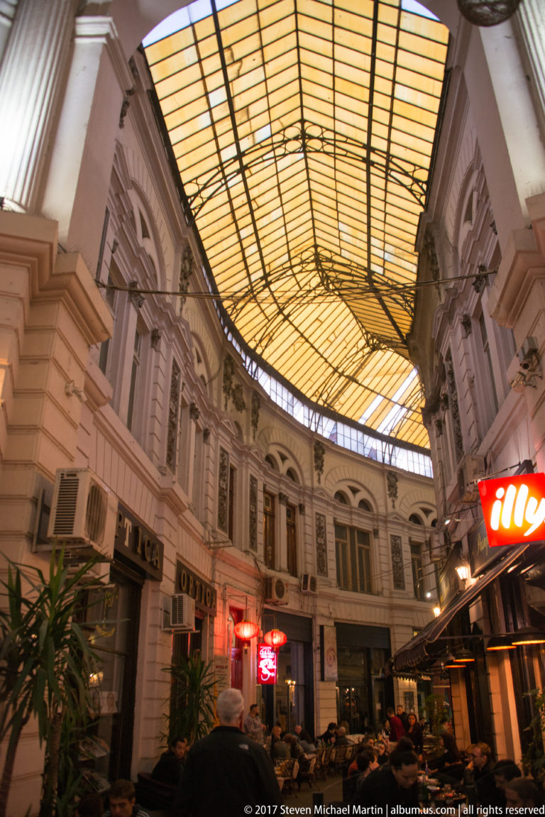 Curved glass-and-metal roof spanning a historic Bucharest arcade, with ornate upper façades and illuminated restaurant signs below.