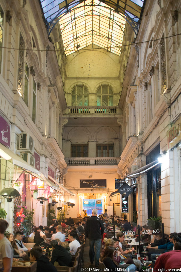 Crowded restaurant tables beneath a high glass roof in a narrow historic Bucharest pedestrian arcade lined with ornate façades.