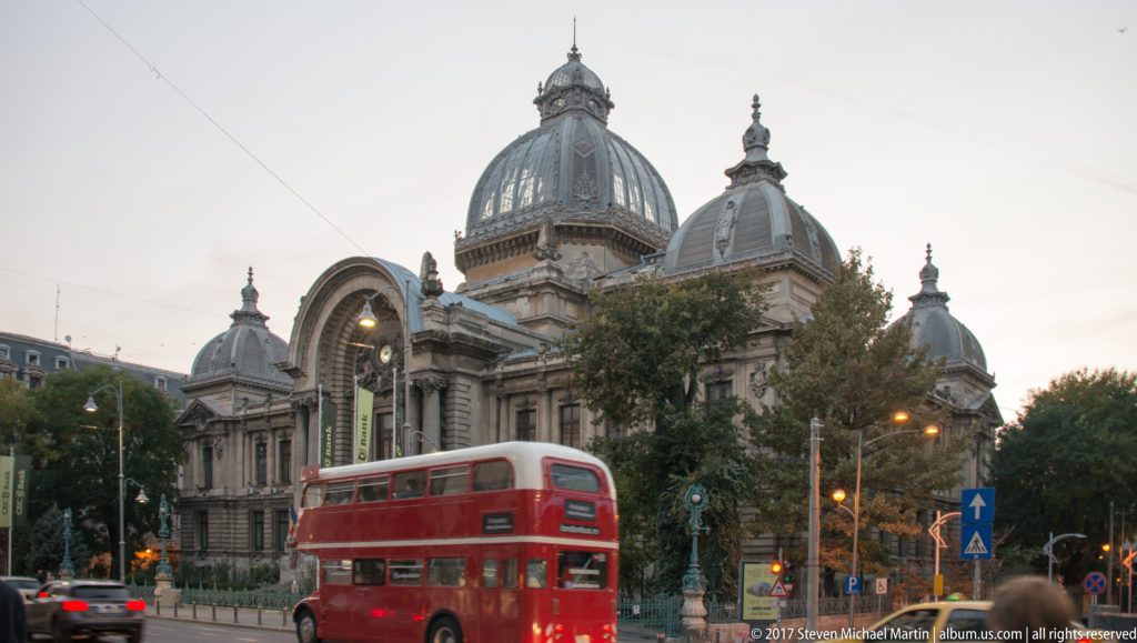 Red double-decker tour bus passing CEC Palace, whose large dome and smaller cupolas rise behind trees at dusk.