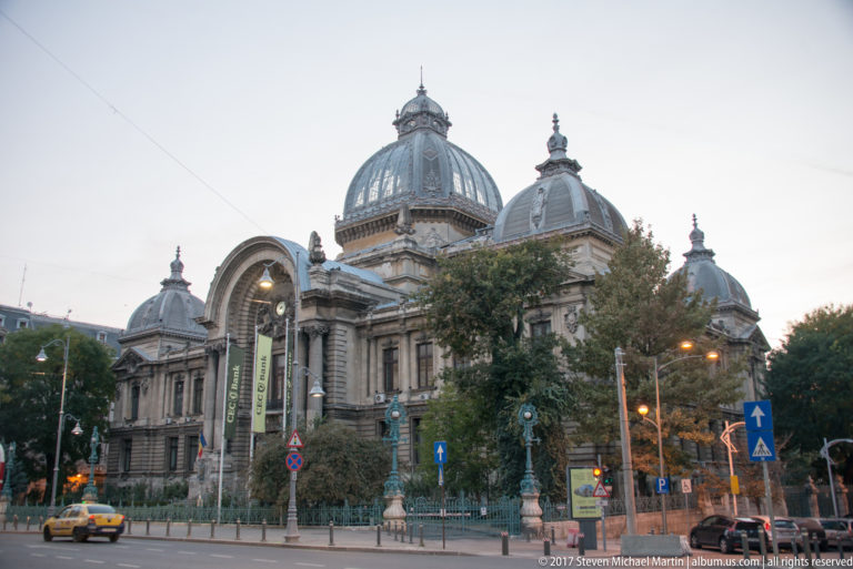 CEC Palace seen across a Bucharest intersection, with its large central dome, smaller cupolas, arched entrance, trees, and street traffic.