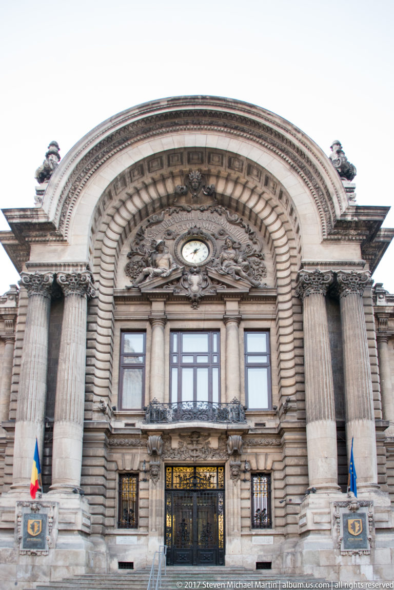 CEC Palace entrance with a huge banded arch, paired columns, clock, carved ornament, gilded doors, and Romanian and EU flags.