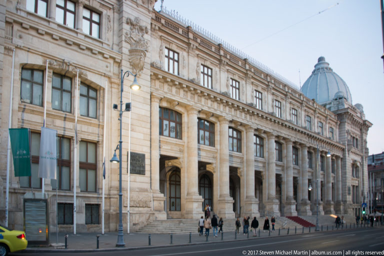 Long historic stone building with arched ground-floor openings and an ornate domed corner pavilion on a Bucharest street.