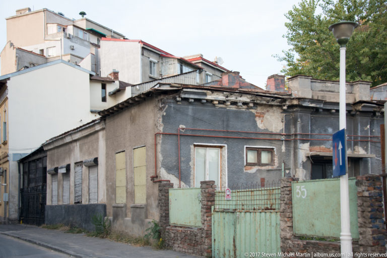 Weathered low-rise Bucharest buildings with exposed brick, patched plaster, metal gates, pipes, and boarded openings.