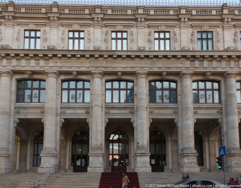 Monumental stone institutional façade with giant columns, sculpted panels, arched entrances, broad steps, and tall windows in Bucharest.