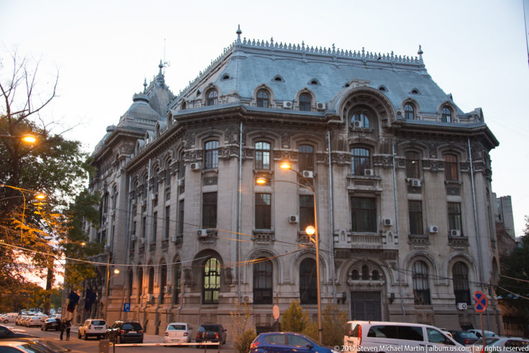 Grand historic corner building with ornate stone façade, mansard roof, dormers, large arched window, and evening streetlights.
