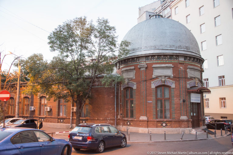 Small round red-brick Bucharest building with gray dome, tall windows, decorative masonry bands, trees, and parked cars.