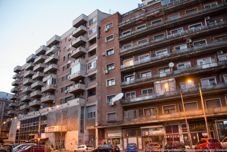 Long brick Bucharest apartment block with balconies, satellite dishes, shops, parked cars, and glowing streetlights at dusk.