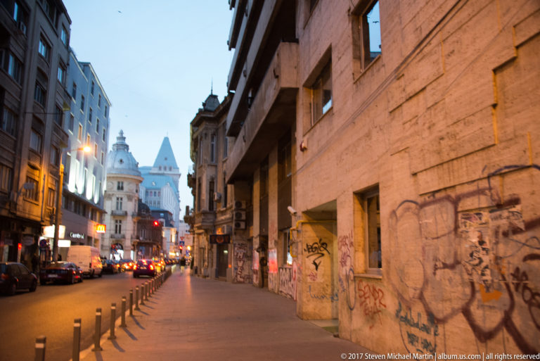 Evening Bucharest street with graffiti-covered wall, cars and bollards leading toward illuminated historic towers in the distance.