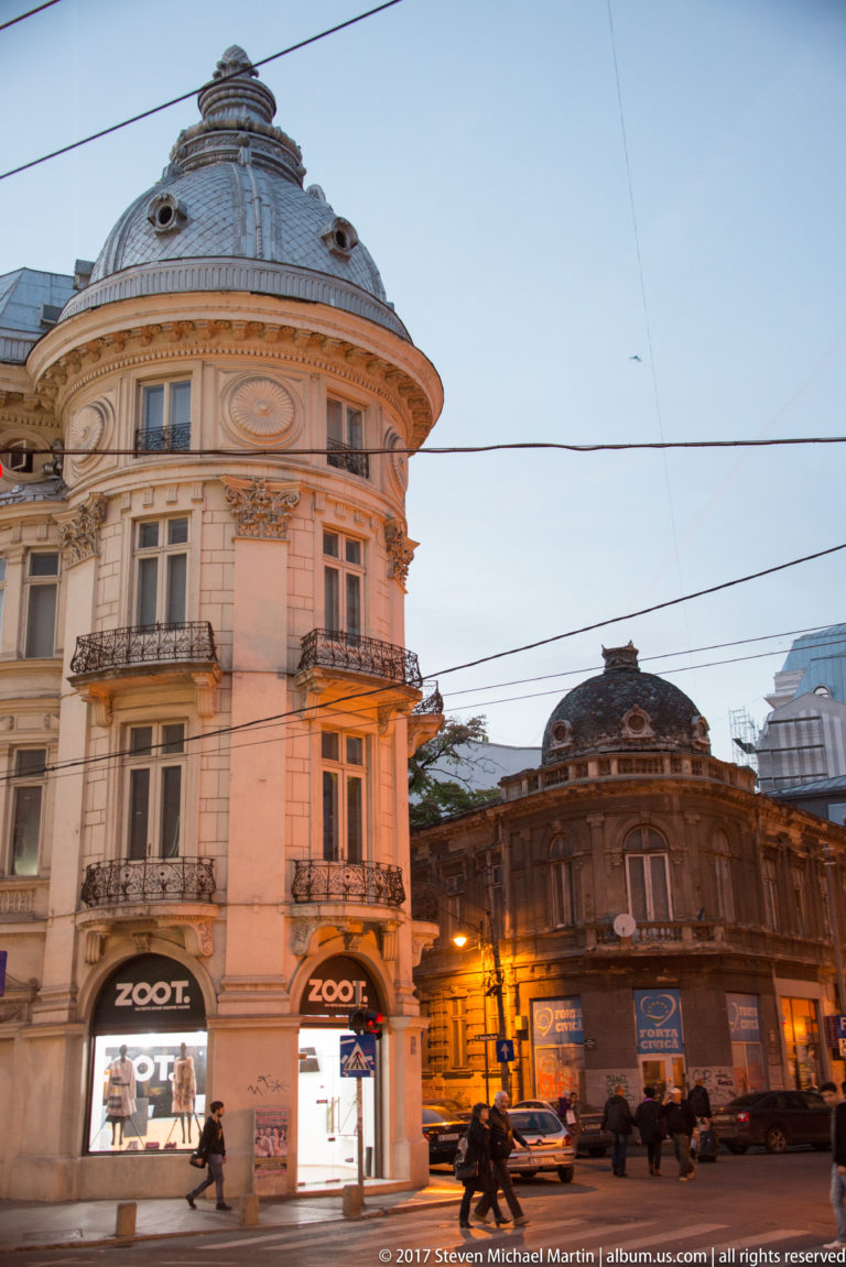 Rounded pale historic corner building with ornate balconies, ZOOT storefronts, overhead wires, and an adjoining lower building at dusk.