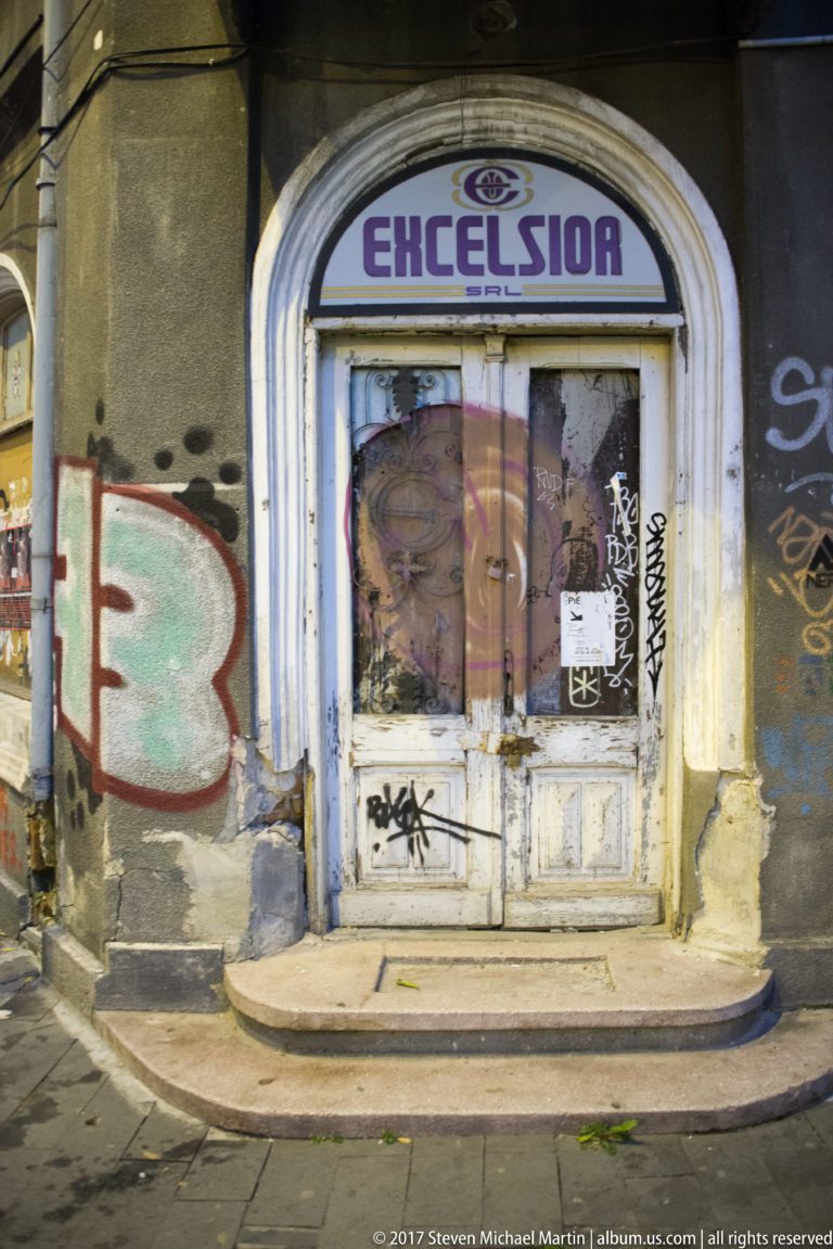 Arched doorway labeled “EXCELSIOR,” with worn double doors, stickers, graffiti, and spray-painted walls on both sides.