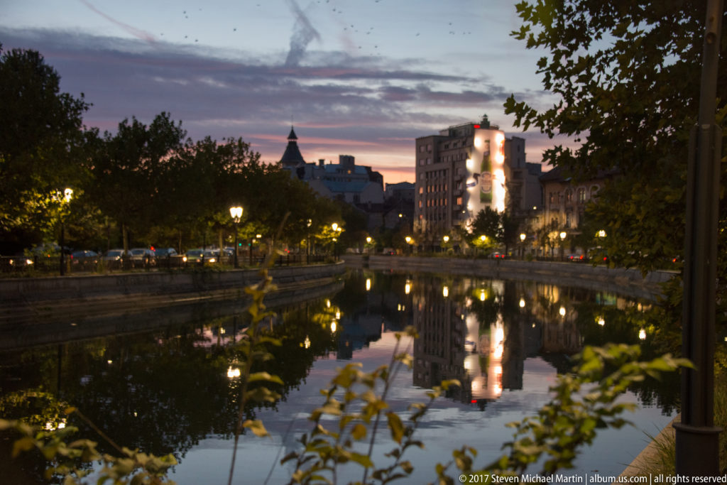 Calm Bucharest lake at dusk reflecting city buildings, glowing streetlamps, trees, and a brightly lit tower.
