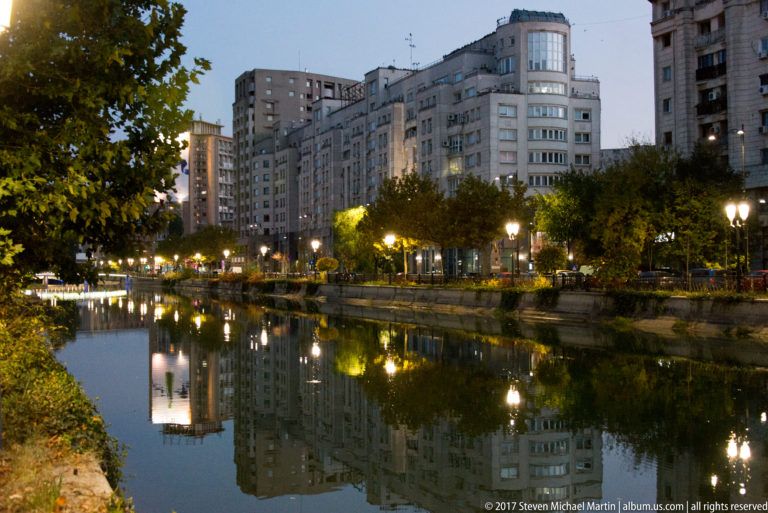 Apartment blocks and trees along a Bucharest lake, reflected in calm water beneath evening streetlamps.
