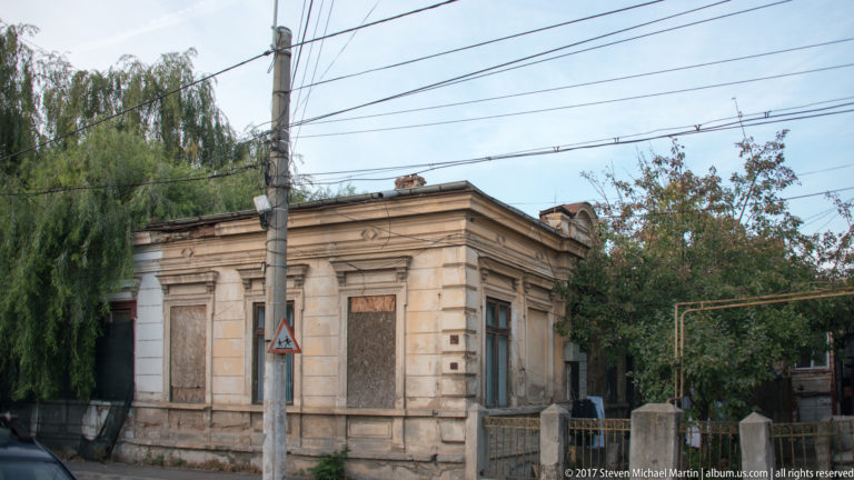 Single-story historic Bucharest house with boarded windows, weathered masonry, low fence, trees, and overhead wires.
