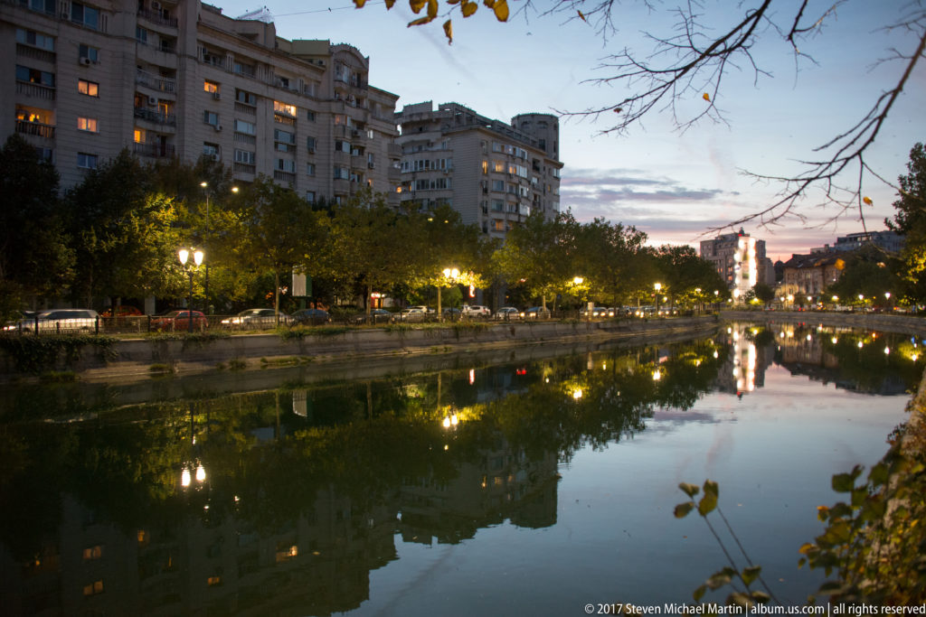 Wide twilight view along a Bucharest lake with apartment blocks, tree-lined shores, glowing lamps, and reflections in still water.