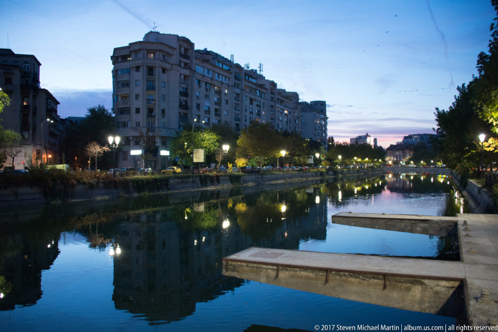 Calm Bucharest river at dusk with apartment blocks, streetlamps, concrete platforms, trees, and reflections in the water.