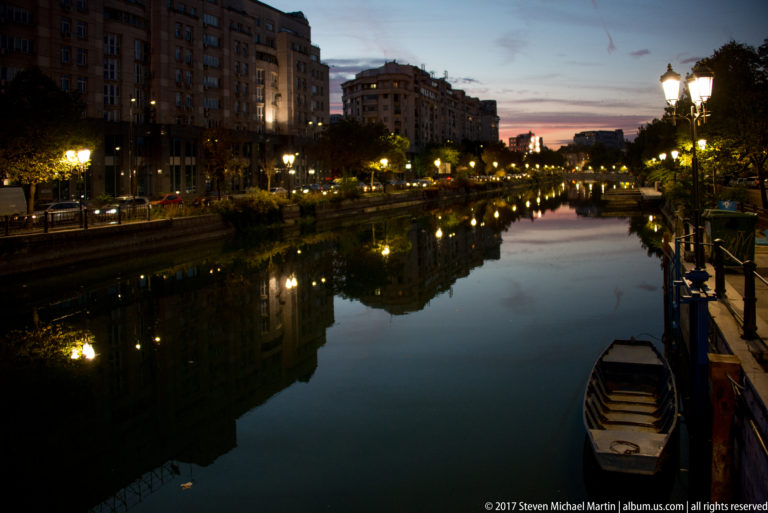 Twilight river in Bucharest with a moored boat, rows of streetlamps, mid-rise buildings, and strong reflections.