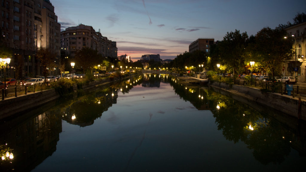 Centered twilight view down a Bucharest river with illuminated embankments, trees, buildings, and mirrored sunset colors.