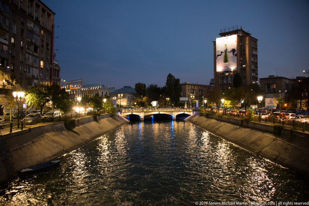 Illuminated bridge over a channelized Bucharest river, with concrete embankments, traffic, trees, and a tall lit building beyond.