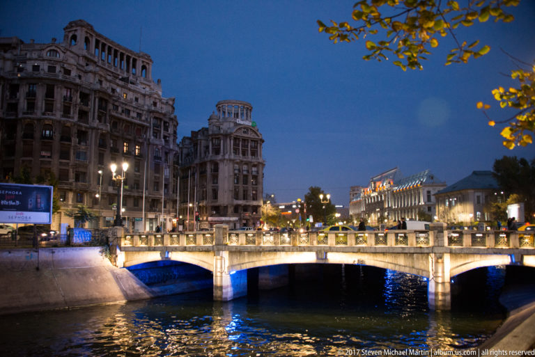 Multi-arch bridge glowing over the Bucharest river, with ornate historic buildings, blue underlighting, and reflected city lights.