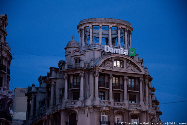 Historic rounded Bucharest building with columns, balconies, circular rooftop pavilion, and illuminated “Damilá” sign.