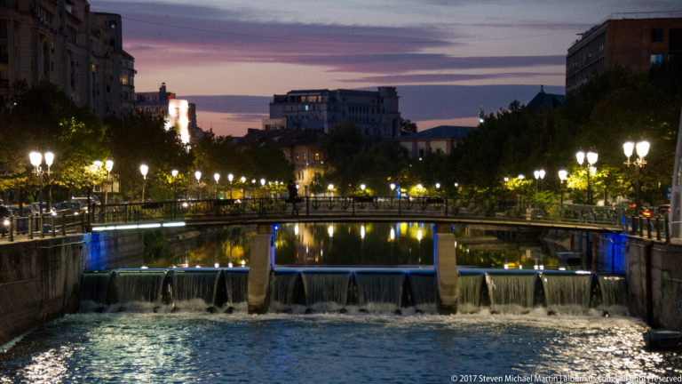 Low weir spilling beneath a pedestrian bridge on a Bucharest river, with lamps, trees, and twilight buildings beyond.