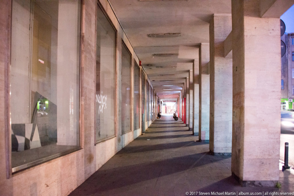 Long covered Bucharest sidewalk arcade with square concrete columns, glass storefronts, ceiling lights, and a distant pedestrian.