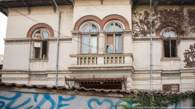 Deteriorated Bucharest façade with paired arched windows, red-brick trim, small balcony, open windows, ivy, and graffiti.